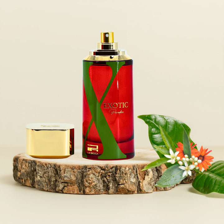 Red perfume bottle with gold cap on a wooden platform, surrounded by green leaves and flowers Rue Broca Exotic Paradise 