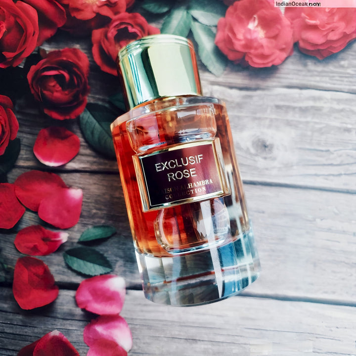 Perfume bottle labeled 'Exclusif Rose' surrounded by red roses and petals on a wooden surface.