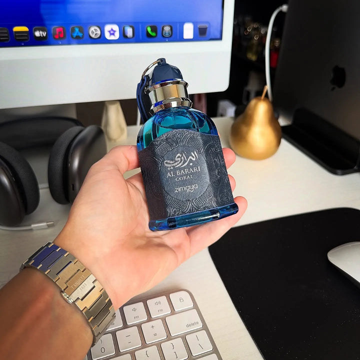 Hand holding a blue perfume bottle with a label in front of a computer monitor on a desk.